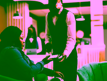 African American woman hotel guest sitting in lobby holding smartphone paying for order using nfc technology. Waiter holding POS terminal for contactless payment serving female traveler